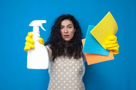 Photo for Young girl cleaner in uniform holds a spray and rags on a blue background, a woman housekeeper in an apron and gloves holds cleaning products - Royalty Free Image