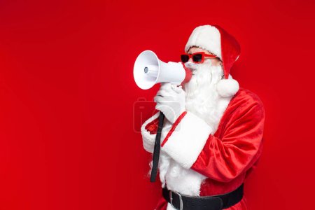 santa claus dans des lunettes rouges annonce des informations en mégaphone sur fond coloré, homme en chapeau et costume de Père Noël pour Noël parle en haut-parleur sur l'espace de copie