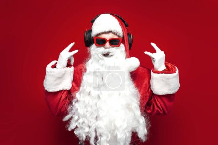 santa claus in hat and festive glasses listens to music in headphones and sings on red background, man in santa costume screams and shows rock gesture with his hands