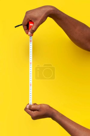 Téléchargez les photos : Mains de l'homme afro-américain tenir mètre ruban et mesurer la distance sur fond jaune isolé, gars avec outil de mesure de construction, gros plan - en image libre de droit