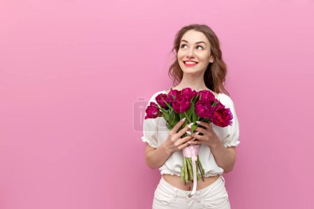 Young attractive girl in white clothes holds bouquet of pink tulips and looks at the copy space, woman on holiday with flowers on pink isolated background