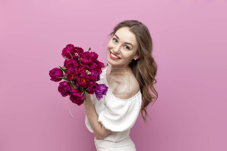 young cute girl in festive clothes holds bouquet of flowers and smiles on pink isolated background, woman with pink tulips celebrates March 8
