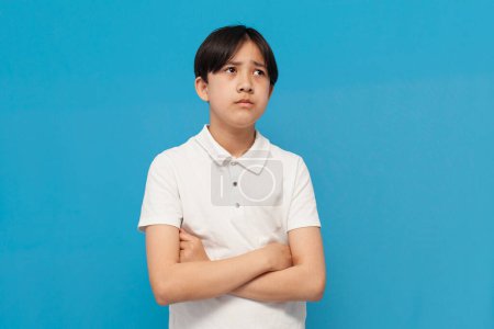 unglücklicher asiatischer Junge von zwölf Jahren ist traurig und beleidigt auf blauem Hintergrund, unzufriedenes koreanisches Kind im weißen T-Shirt steht mit verschränkten Armen und denkt