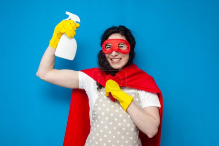 Photo for Young girl cleaner in gloves and superman costume holds detergent on blue background, woman housewife in superhero mask shows cleaning spray - Royalty Free Image
