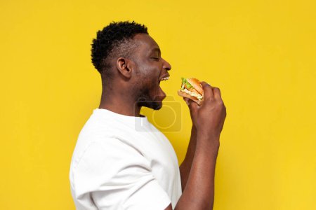Photo for African american man in white t-shirt bites big burger on yellow isolated background, young guy eats fast food, side view - Royalty Free Image