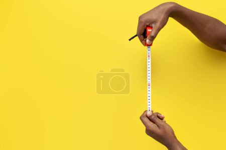 Téléchargez les photos : Mains de l'homme afro-américain tenir mètre ruban sur fond jaune isolé, les mains mesurent la distance et la taille avec du ruban adhésif, gros plan - en image libre de droit