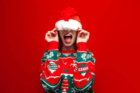 Téléchargez les photos : Mignonne fille en pull de Noël se cache dans un grand chapeau de Père Noël et crie sur fond rouge, femme embarrassée dans les vêtements de Noël ferme les yeux avec un chapeau et ouvre la bouche - en image libre de droit
