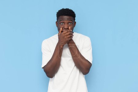 schockierter junger Mann Afroamerikaner in weißem T-Shirt verdeckt seinen Mund mit den Händen auf blauem Hintergrund, verängstigter Mann zeigt sich überrascht und erstaunt