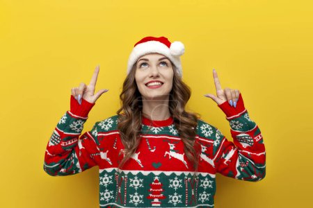 Photo for Young girl in christmas sweater and santa claus hat smiles and points up at copy space on yellow isolated background, woman in christmas clothes advertises empty space from above - Royalty Free Image