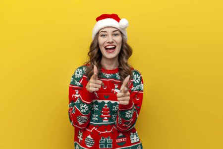 Photo for I choose you. young girl in christmas sweater and santa claus hat smiles and points at the camera on yellow isolated background, woman in christmas clothes points forward with her hands - Royalty Free Image