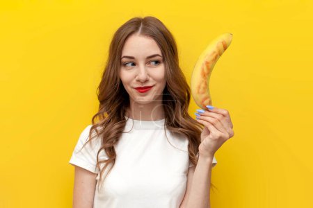 young girl shows kissed banana and hints at intimacy on yellow isolated background, woman with fruit smiles, sexy and erotic concept