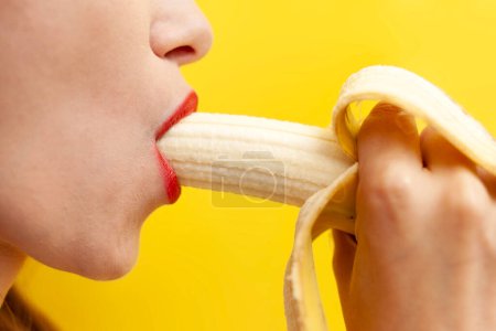 sexy and intimate blowjob concept, woman licks and takes banana in her mouth on yellow background, the girl's tongue and lips erotically touch the fruit