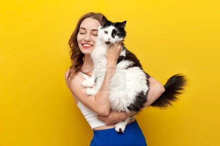 Photo for Young cute girl holding black and white cat on yellow isolated background and smiling, woman with pet posing on colored background, care and love for animals - Royalty Free Image