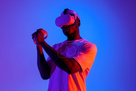 Foto de Hombre afroamericano en gafas de realidad virtual apuntando y disparando en iluminación de neón, chico gamer jugando y luchando en gafas vr con joysticks - Imagen libre de derechos
