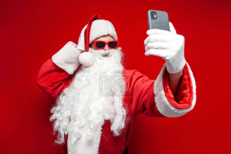 Weihnachtsmann mit Hut und festlicher Brille nutzt Smartphone und macht Selfie auf rotem Hintergrund, Mann im Weihnachtsmannkostüm spricht via Videolink online
