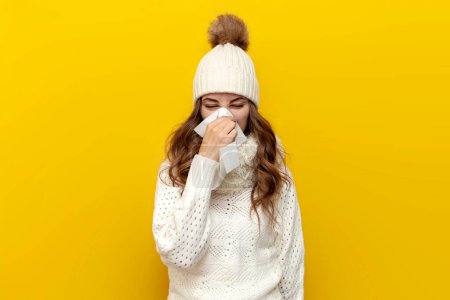 sick girl in warm soft winter clothes with runny nose in napkin on yellow isolated background, cold woman in white hat and scarf in comfortable and cozy knitted sweater shows flu and cold symptoms