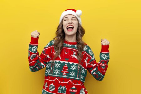 Photo for Young girl in christmas sweater and santa claus hat celebrates victory and success on yellow isolated background, woman winner rejoices in luck in new year's clothes - Royalty Free Image