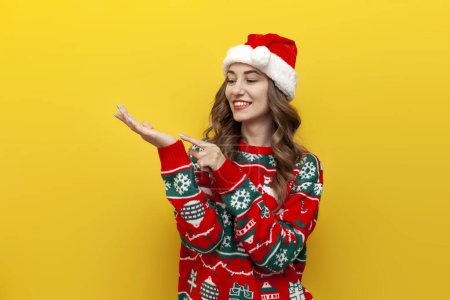 Photo for Young girl in christmas sweater and santa claus hat holds empty hands in front of her on yellow isolated background, woman advertises copy space in new year's clothes - Royalty Free Image