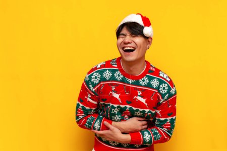 Foto de Joven asiático hombre en navidad suéter y santa hat risas y se burla de en amarillo aislado fondo, coreano chico en nuevo año ropa chistes y espectáculos ridiculizar - Imagen libre de derechos