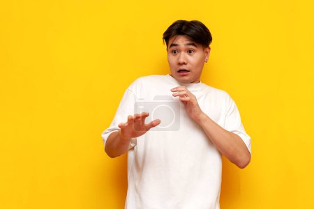 shocked scared young asian guy afraid and covering himself with hands on yellow isolated background, shocked korean man in white t-shirt in amazement in panic