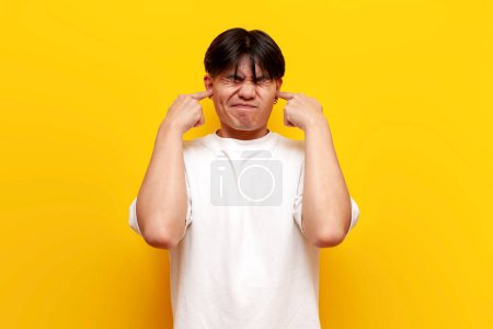 Foto de Joven asiático chico en blanco camiseta cubierto su oídos con su manos de ruidoso sonido y evita el ruido en amarillo aislado fondo, coreano hombre ignora y no escucha - Imagen libre de derechos