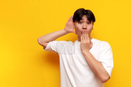 Foto de Sorprendido joven asiático chico en blanco camiseta escuchando secreto información sobre amarillo aislado fondo coreano hombre espiando y sorprendido - Imagen libre de derechos