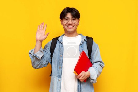 Téléchargez les photos : Jeune garçon asiatique étudiant en denim chemise et lunettes avec sac à dos tenant des livres et saluant sur fond jaune isolé, coréen homme saluant et disant au revoir - en image libre de droit