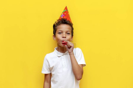 African American boy in festive hat celebrates his birthday and blows trumpet on blue isolated background, 10 year old child rejoices and makes noise for the holiday