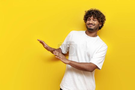 Téléchargez les photos : Jeune homme indien évitant et refusant sur fond jaune isolé, mécontent homme sud-asiatique en t-shirt blanc ignorant et montrant dégoût - en image libre de droit