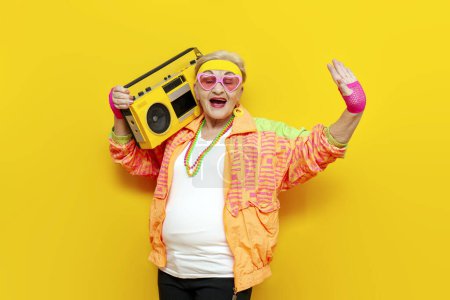 Foto de Divertido viejo abuelita con grabadora en deportes hipster ropa toma selfie en línea en amarillo aislado fondo, anciana en elegante ropa juvenil escucha música fuerte y baila en la discoteca - Imagen libre de derechos