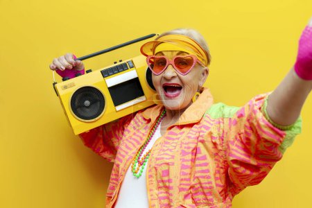 Foto de Divertida abuela vieja loca con grabadora en ropa hipster deportes escucha música y canta sobre fondo aislado amarillo, anciana en ropa de juventud con estilo con tocadiscos grita en la fiesta - Imagen libre de derechos