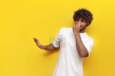 Téléchargez les photos : Jeune bouclé indien homme montrant dégoût et couvrant son nez avec la main sur fond jaune isolé, gars refuse mauvaise odeur et rejette la puanteur - en image libre de droit