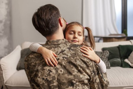 Photo for Young Ukrainian soldier in a camouflage uniform hugs a child at home and says goodbye, a little girl hugs and greets a military dad, a happy child meets his father from the army - Royalty Free Image