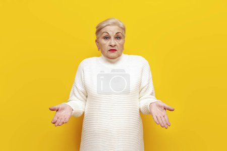 unsure old grandmother in a white sweater shrugs her shoulders puzzled over yellow isolated background, confused elderly woman pensioner spreads her arms and doesnt know