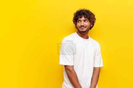 junger bescheidener indischer Mann in weißem T-Shirt schüchtern auf gelbem Hintergrund, ein unsicherer Typ denkt nach und träumt