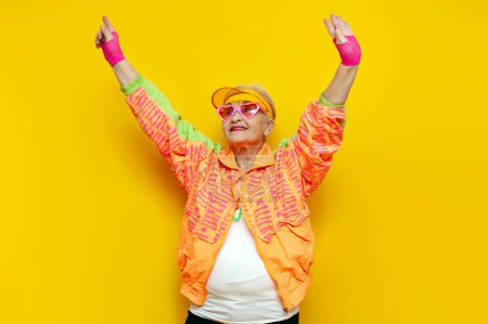 fou drôle vieille mamie dans le sport coloré vêtements et lunettes roses dansant sur un fond jaune isolé, femme âgée dans la jeunesse vêtements hipster se déplaçant avec les mains levées