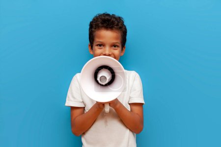 Niño afroamericano en polo blanco anuncia información en megáfono sobre fondo azul aislado, niño de 10 años grita en altavoz