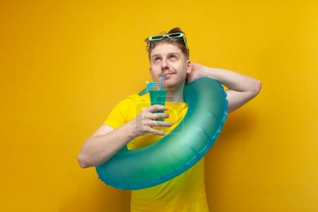 junger Kerl im Sommer im Urlaub mit schwimmendem aufblasbarem Ring, der einen erfrischenden blauen Cocktail trinkt und vor gelbem Hintergrund träumt, ein Mann mit einem Sommerdrink, der nachdenkt und aufblickt