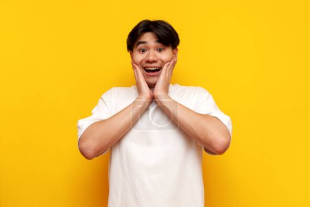 shocked asian young man in white t-shirt surprised with open mouth over yellow isolated background, excited korean guy student screaming in amazement