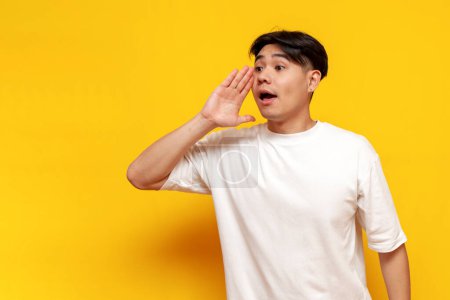 asiatischer junger männlicher Student in weißem T-Shirt kündigt Informationen auf gelbem Hintergrund an, koreanischer Typ schreit und spricht laut