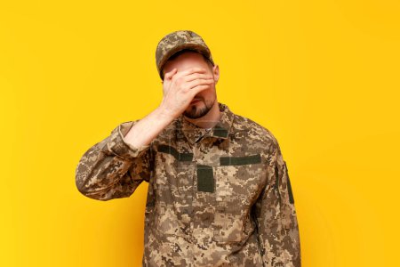 young Ukrainian army soldier in camouflage pixel uniform shows facepalm and failure gesture on yellow isolated background, Ukrainian military cadet fails