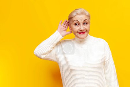 cheerful old granny in a white sweater eavesdropping on secret information on a yellow isolated background, elderly pensioner woman holding her hand near her ear and listening