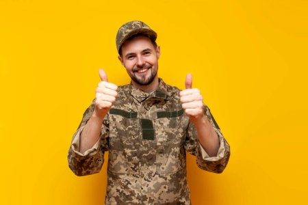 young Ukrainian army soldier in camouflage pixel uniform shows likes with hands and recommends on yellow isolated background, Ukrainian military cadet approves and shows thumbs up