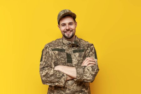 young Ukrainian army soldier in pixel camouflage uniform stands with his arms crossed on a yellow isolated background, Ukrainian military cadet looks at the camera and smiles