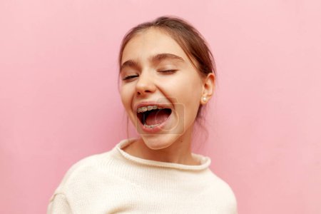 fröhliches Teenie-Mädchen mit Zahnspange zwinkert und deutet auf rosa Hintergrund, fröhliches Kind blinzelt mit den Augen und scherzt