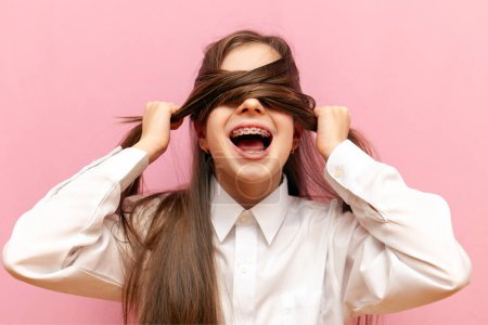 ängstliches kleines Teenager-Mädchen mit Zahnspange schließt die Augen und schreit auf rosa isoliertem Hintergrund, Kind im Stress zieht Haare aus und versteckt sich