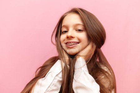Téléchargez les photos : Petite adolescente avec bretelles souriantes et tenant les cheveux longs sur fond rose isolé, enfant regardant la caméra et tenant la tête avec les mains - en image libre de droit