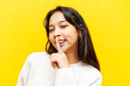 junge fröhliche asiatische Frau mit Zahnspange zeigt Finger in Lippennähe und Geste des Geheimnisses auf gelbem isolierten Hintergrund, koreanisches Mädchen schweigt und verbietet zu sprechen
