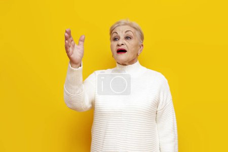 joyeuse vieille grand-mère dans un pull blanc annonce et parle sur un fond jaune isolé, femmes âgées retraité appelle et crie