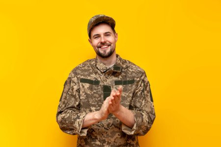 young Ukrainian army soldier in camouflage pixel uniform looks at the camera and applauds on a yellow isolated background, Ukrainian military cadet smiles and claps his hands